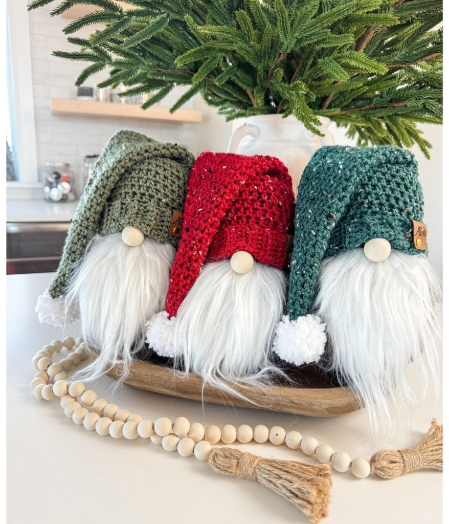 Three crochet gnomes with long white beards and knitted hats in green, red, and teal sit on a wooden tray next to a beaded garland, in front of a small fir tree in a vase.