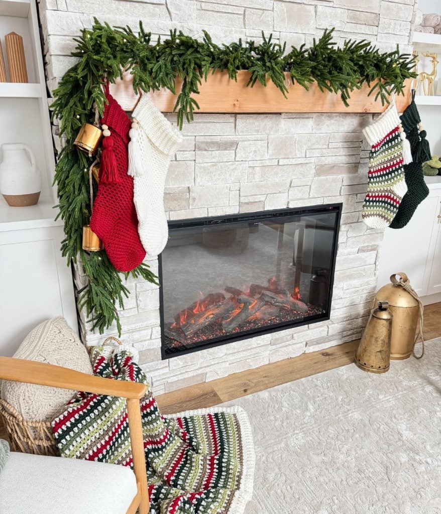 A cozy living room with a fireplace, adorned with a garland and crochet stockings. A striped blanket from the Holiday Weave collection drapes over a chair, while a metallic lantern sits nearby on a rug.