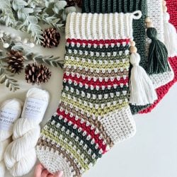 Knitted holiday stockings in green, red, and beige with tassels are displayed alongside yarn skeins, pine cones, and greenery, creating a cozy atmosphere perfect for showcasing a crochet stocking or exploring a free pattern for your own festive creation.