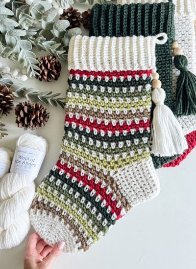 Knitted holiday stockings in green, red, and beige with tassels are displayed alongside yarn skeins, pine cones, and greenery, creating a cozy atmosphere perfect for showcasing a crochet stocking or exploring a free pattern for your own festive creation.