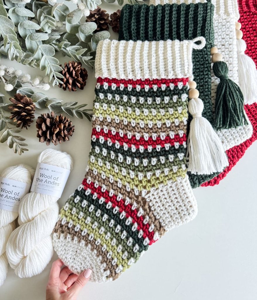 Knitted holiday stockings in green, red, and beige with tassels are displayed alongside yarn skeins, pine cones, and greenery, creating a cozy atmosphere perfect for showcasing a crochet stocking or exploring a free pattern for your own festive creation.