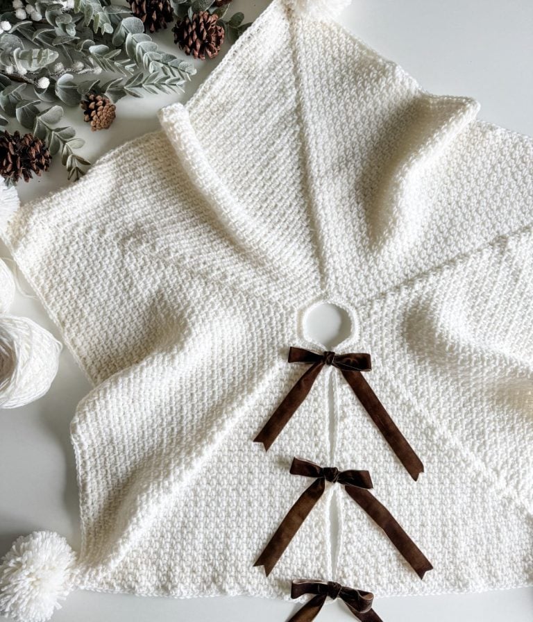 White textured knit poncho with a hood, brown ribbon ties, and decorative pom-poms, evokes holiday charm as it lays surrounded by pinecones and greenery—a design reminiscent of a festive tree skirt.
