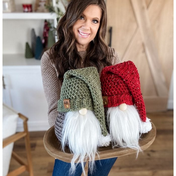 In a cozy room with wooden flooring and white shelves, a person holds a wooden tray with two crochet gnome decorations, one in green and one in red.