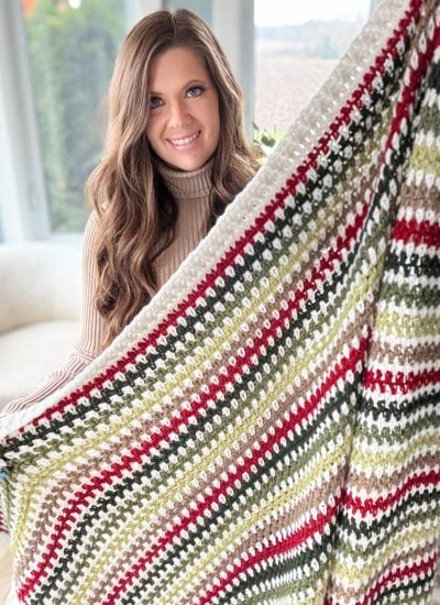 A person proudly displays a vibrant crocheted blanket featuring green, red, and white stripes, showcasing an intricate crochet pattern that adds to its charm.