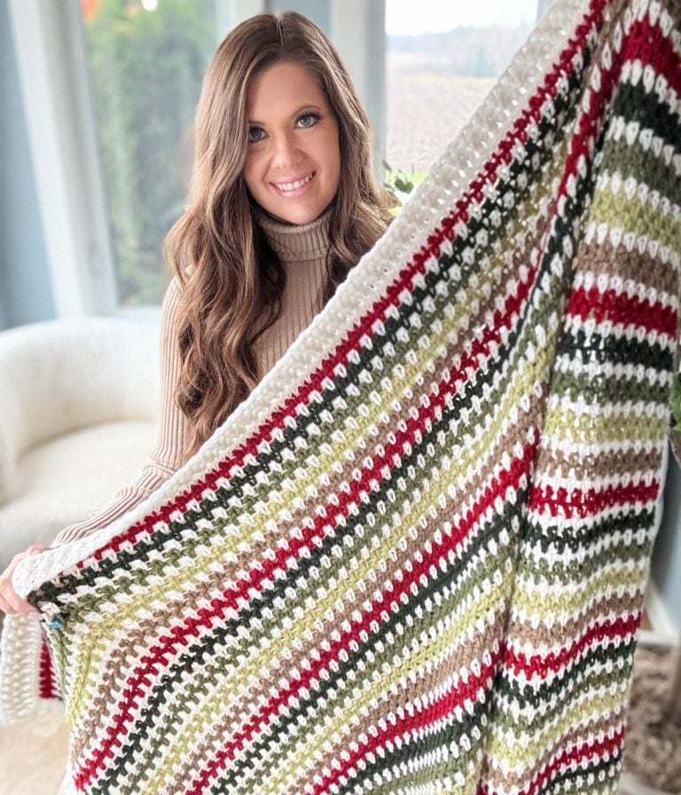 A person proudly displays a vibrant crocheted blanket featuring green, red, and white stripes, showcasing an intricate crochet pattern that adds to its charm.