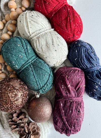 A charming crochet gnome sits among various colored yarn balls, nestled with Christmas ornaments and pinecones on a textured surface.
