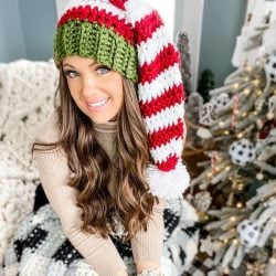 A woman with long hair wears a red and white striped crochet elf hat with a green band, sitting indoors beside a decorated Christmas tree.