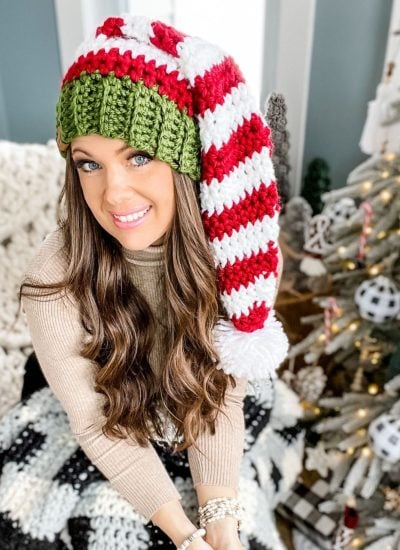 A woman with long hair wears a red and white striped crochet elf hat with a green band, sitting indoors beside a decorated Christmas tree.