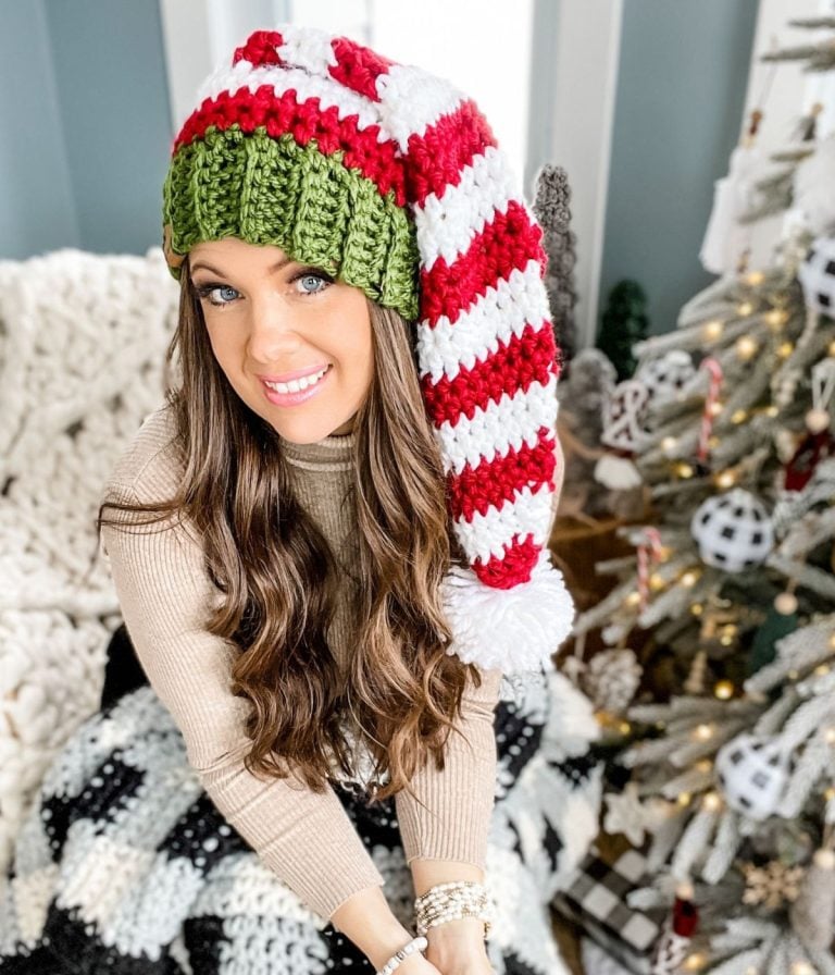 A woman with long hair wears a red and white striped crochet elf hat with a green band, sitting indoors beside a decorated Christmas tree.