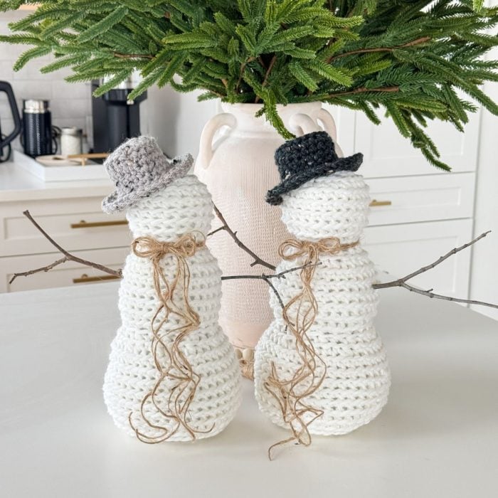 Two rustic farmhouse snowmen with twig arms and hats stand on a white surface, complemented by a green plant in the background.