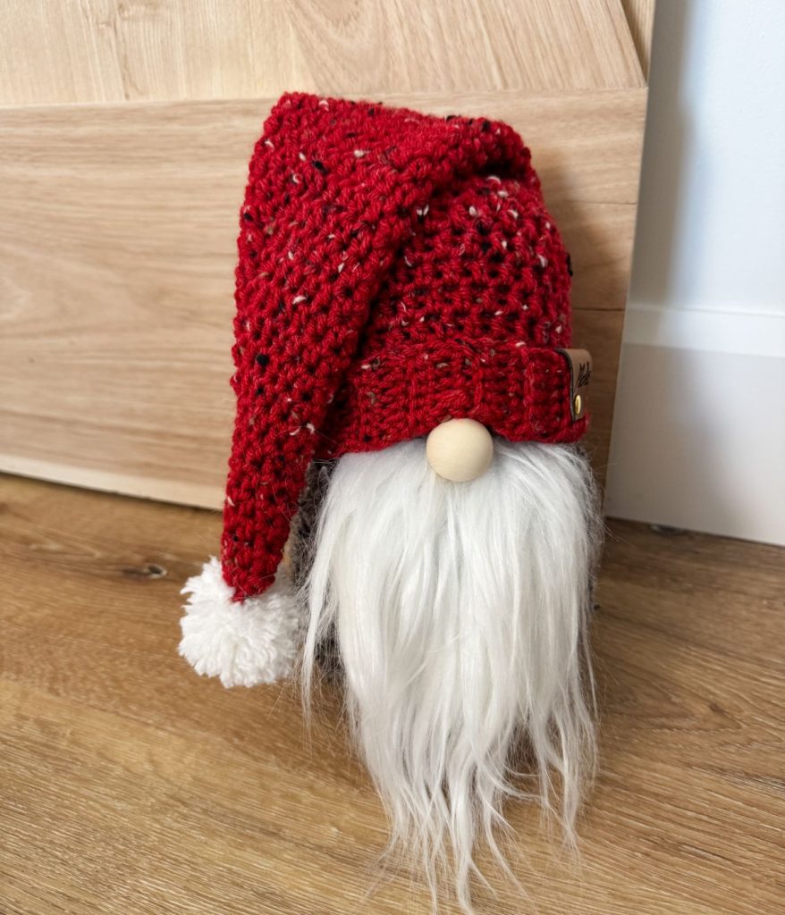 A crochet gnome with a long white beard and a red knitted hat stands proudly on a wooden floor.
