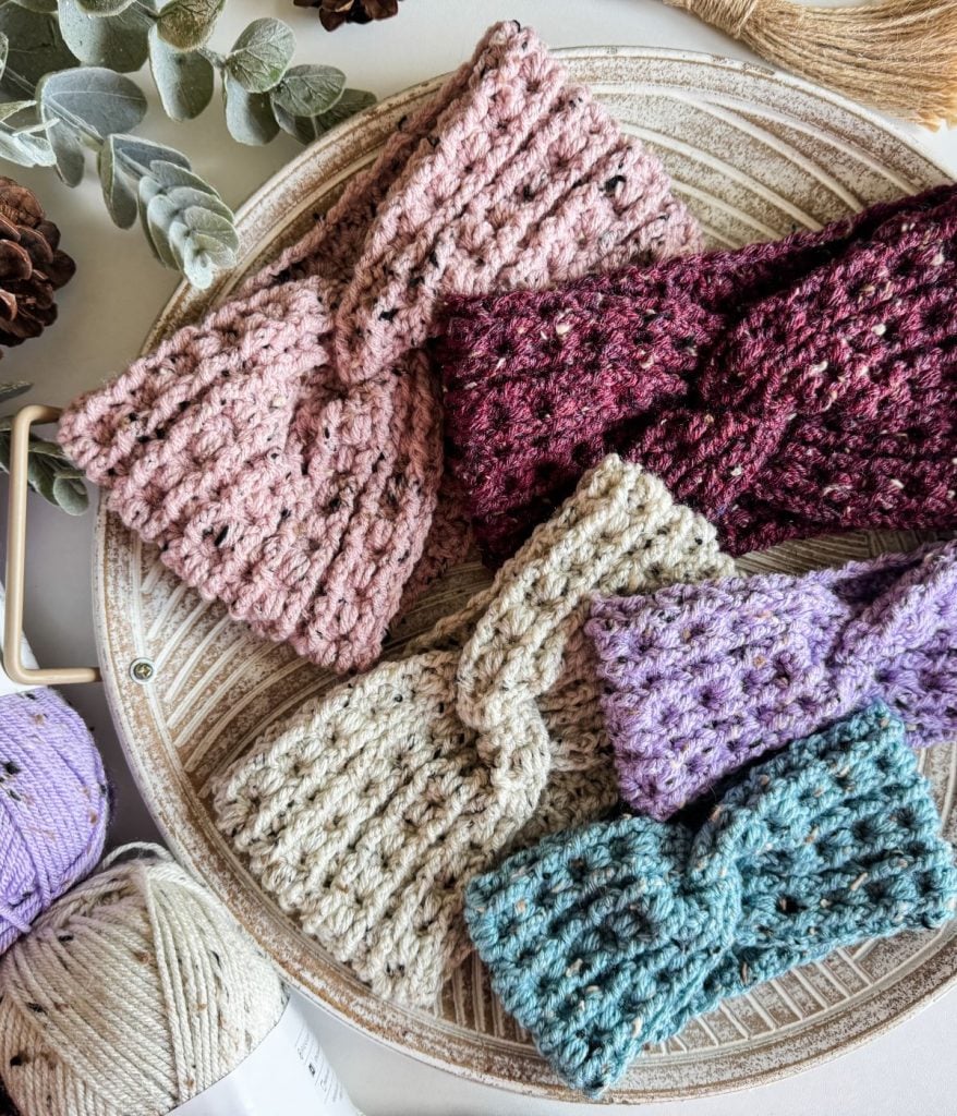 A round tray elegantly displays a collection of crochet headbands in shades of pink, maroon, beige, purple, and teal. Surrounding the array are artfully arranged yarn and eucalyptus leaves, adding a touch of nature's charm.