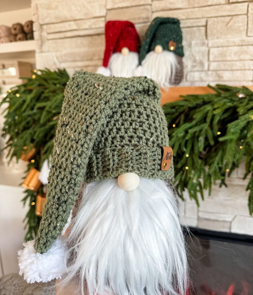 A charming crochet gnome with a green hat and white beard stands in the foreground, surrounded by fellow gnomes wearing red and green hats atop a tastefully decorated mantle.