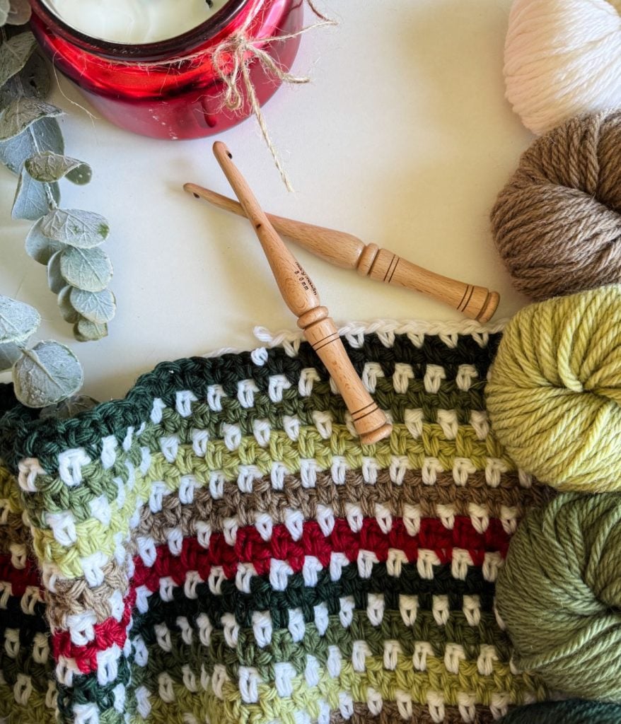 A crocheted fabric with green, red, and white stripes crafted into a cozy blanket is surrounded by yarn balls, two wooden crochet hooks, eucalyptus leaves, and a red candle. The intricate design showcases the moss stitch, adding texture to this charming crochet pattern setting.
