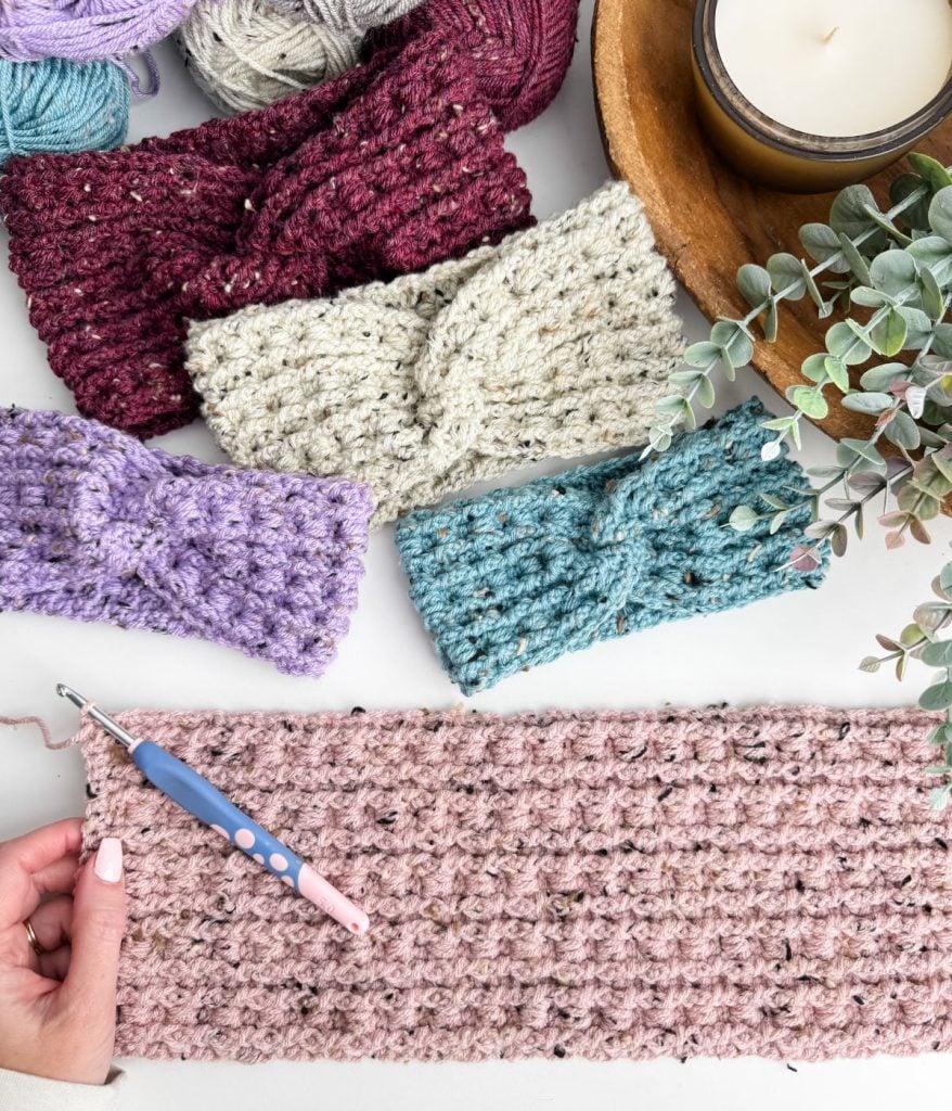 A hand holds a crochet hook next to a pink crocheted piece. Nearby, vibrant crochet headbands in red, white, purple, and teal are displayed alongside yarn and a candle on the table.