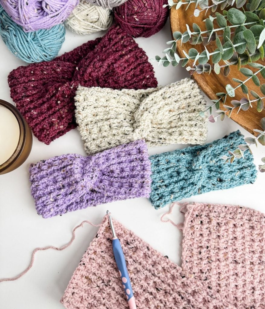 Crocheted headbands in various colors, a crochet hook, and a partially completed crochet headband project are artfully displayed on a white surface alongside colorful yarn.