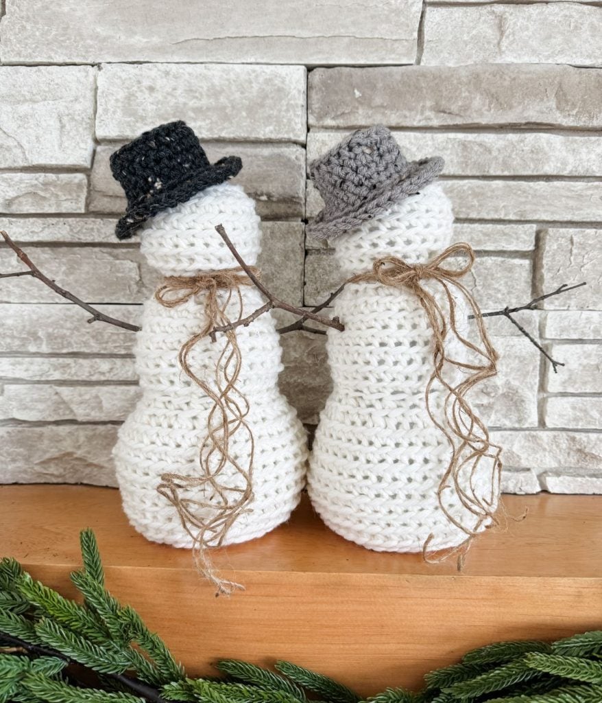 Two crocheted snowmen, crafted from a free crochet pattern, don black and gray hats, twig arms, and yarn scarves. They sit charmingly on a wooden surface against a stone background, embodying the essence of rustic farmhouse decor.
