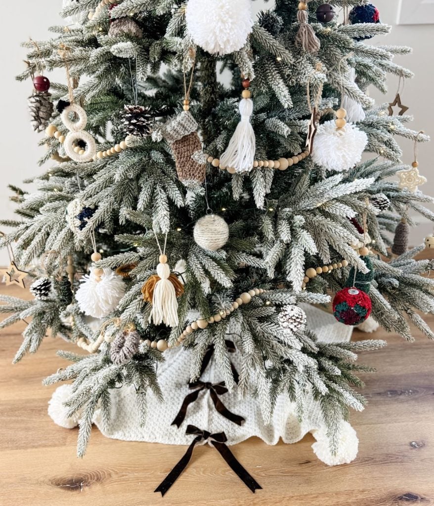 A decorated Christmas tree with handmade ornaments, including yarn pom-poms, pine cones, and wood beads, stands on a wooden floor adorned with a crochet tree skirt.