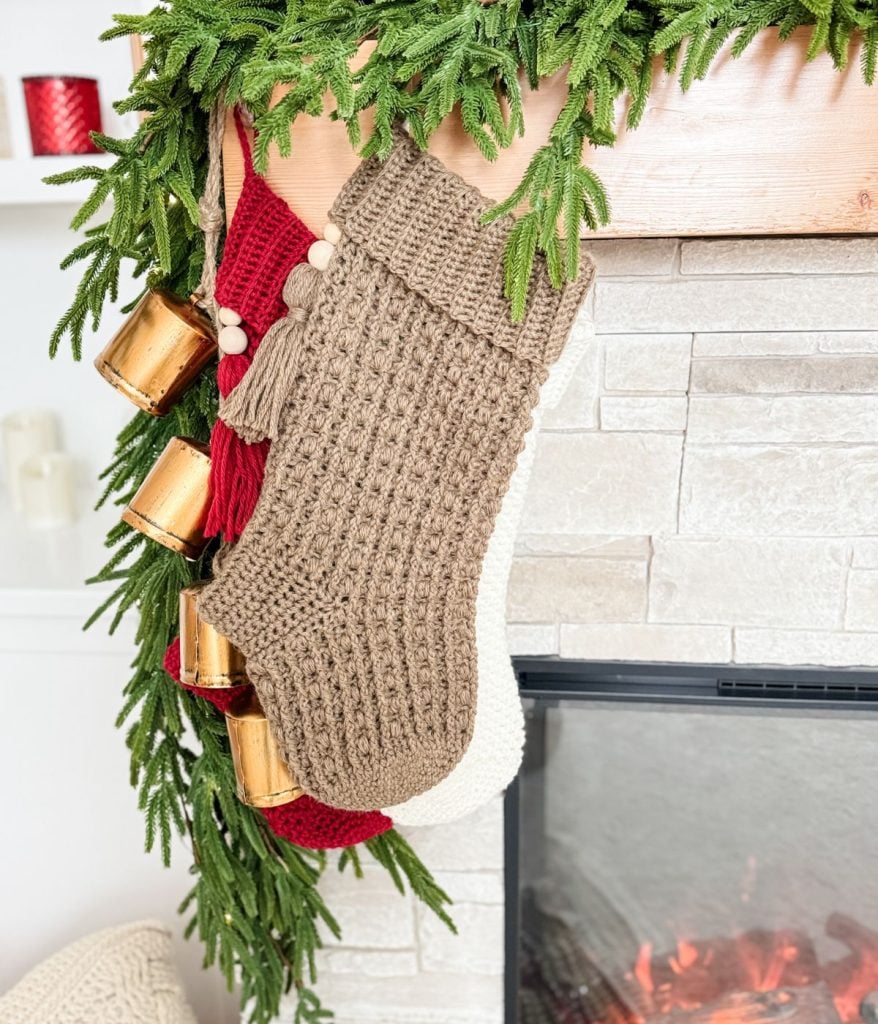 Three knitted stockings hang from a fireplace mantel adorned with green garland and golden bells, capturing the cozy charm of Snuggle Season Stockings.
