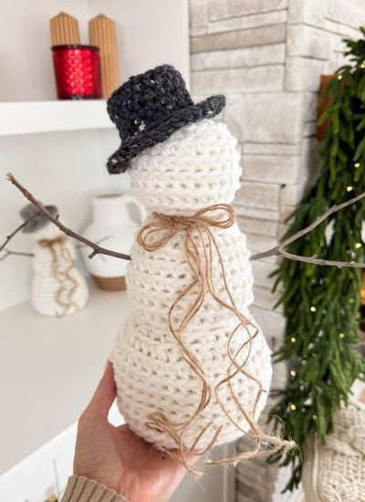 A hand holding a small Rustic Farmhouse Snowman, complete with a black hat and twig arms, in a room adorned with greenery and a red candle.