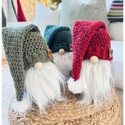 Three charming gnomes—two knitted and one crochet—sit with their long white beards and red and green hats on a woven stool.