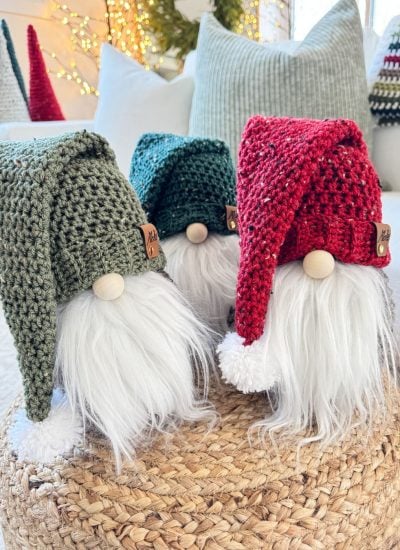 Three charming gnomes—two knitted and one crochet—sit with their long white beards and red and green hats on a woven stool.