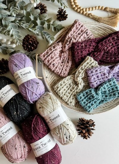 A variety of knitted and crochet headbands are displayed on a round woven mat. Balls of Brava Tweed yarn in assorted colors are arranged nearby, surrounded by pinecones and eucalyptus leaves.