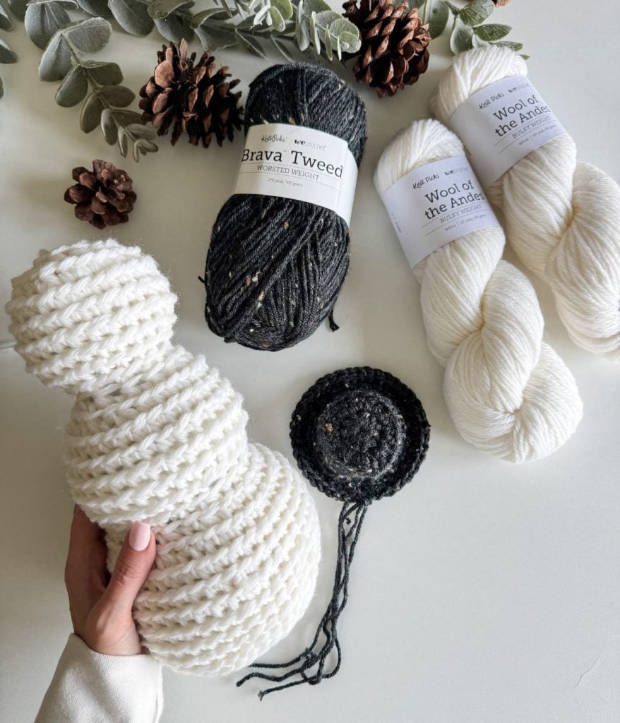 A hand holds a partially knitted white snowman figure, crafted in a rustic farmhouse style, surrounded by yarn balls and skeins on a white surface. Pine cones and eucalyptus leaves add natural charm to this cozy scene.