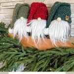 Three charming crochet gnomes with white beards and festive green and red hats grace the mantel alongside lush greenery. Text reads: "Rustic Farmhouse Holiday Gnome - Crochet Pattern & Video".