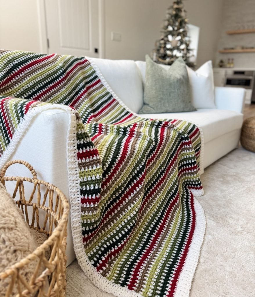 A white sofa with a green, red, and white striped crochet blanket featuring a delightful moss stitch pattern is draped over it. A Christmas tree stands merrily in the background.