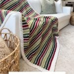 A striped crochet blanket, crafted with a holiday weave, is elegantly draped over a white sofa in a cozy living room, with a festive Christmas tree glowing in the background.
