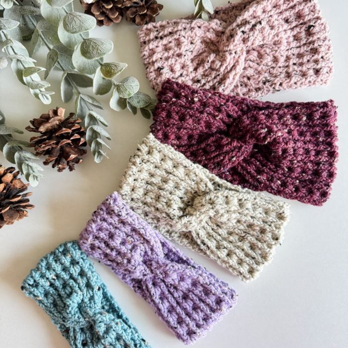 Four knitted and crochet headbands in pink, burgundy, cream, and blue are elegantly displayed on a surface with pine cones and eucalyptus leaves nearby.