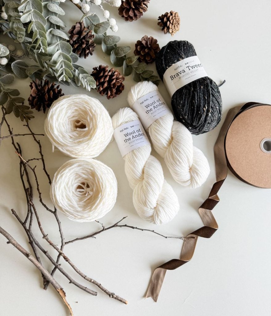 Yarn balls and skeins with labels, surrounded by pine cones, foliage, twigs, and ribbons on a white background evoke a charming rustic farmhouse vibe. Perfect inspiration for your next free crochet pattern.