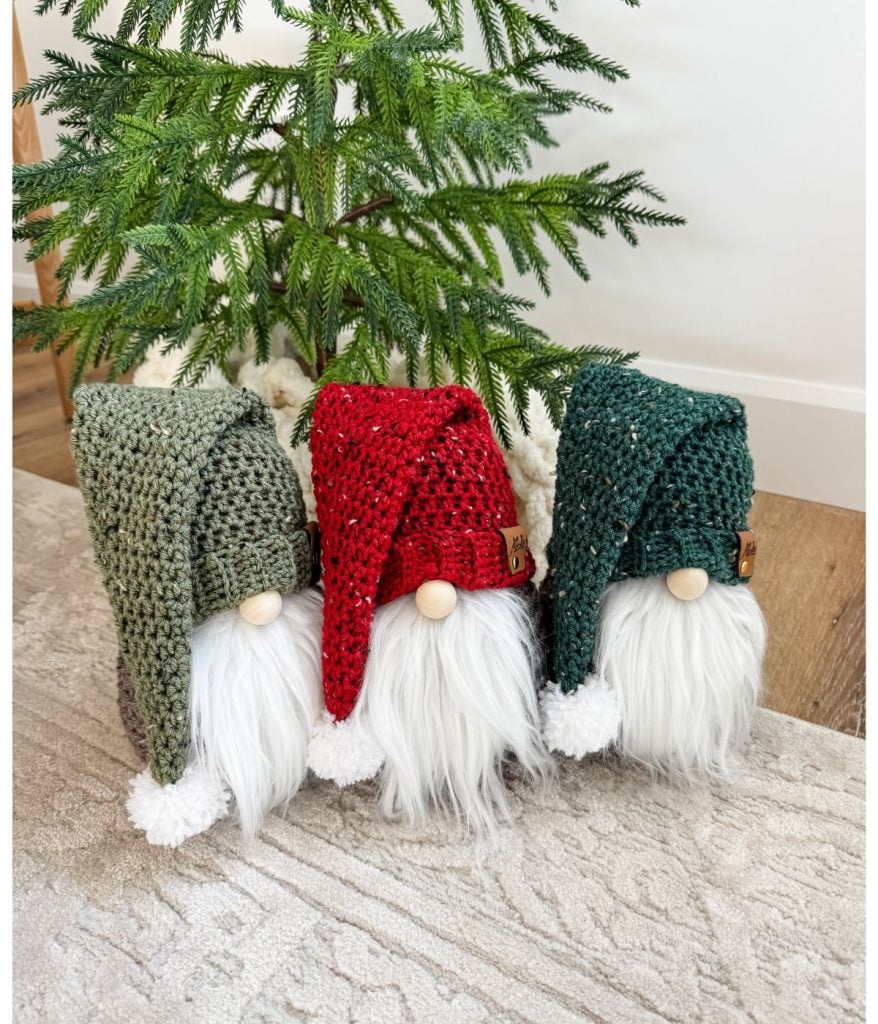 Three crochet gnome figures with long white beards and knitted hats in green, red, and dark green sit cozily in front of a small evergreen tree on a patterned rug.