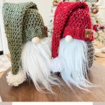 Two handmade crochet gnomes with long white beards and large knitted hats in green and red, posed in front of a decorated Christmas tree.