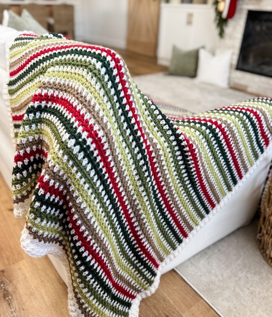 A crocheted blanket featuring Moss Stitch with green, red, white, and beige stripes draped over a white sofa in a living room.