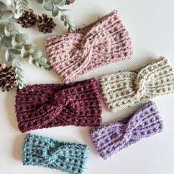 Five knitted and crochet headbands in pink, beige, maroon, purple, and teal are laid out on a white surface, surrounded by greenery and pinecones.