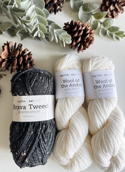 Yarn skeins, featuring two white "Wool of the Andes" and one dark "Brava Tweed," sit amidst pinecones and foliage, evoking a rustic farmhouse snowman vibe.