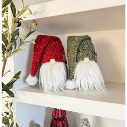 Two decorative crochet gnomes with long white beards and knitted hats, one red and one green, are displayed on a shelf surrounded by small holiday tree decorations nearby.