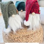 Three crocheted holiday gnomes with red and green hats sit on a woven surface. A sign below reads, "Rustic Farmhouse Crochet Gnome Pattern & Video.