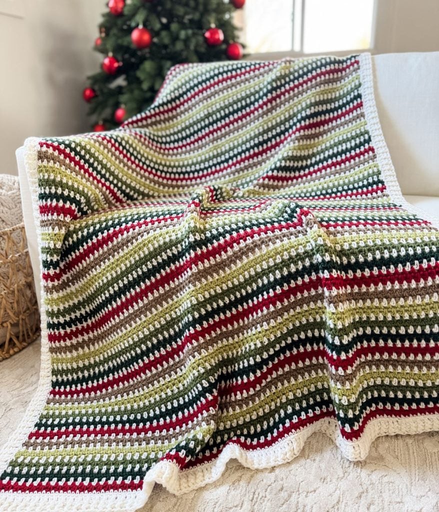 A beautifully crocheted blanket featuring a moss stitch pattern in red, green, and beige stripes is draped over a white sofa. In the background, a Christmas tree adorned with red ornaments completes the cozy setting.