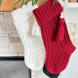 Two knitted Christmas stockings, one white and one red, hang from a mantel adorned with greenery—perfect inspiration for those seeking a Snuggle Season Stocking or a crochet stocking pattern to cozy up their holiday decor.