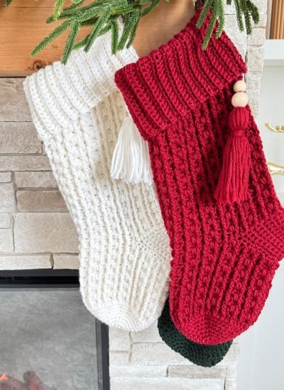 Two knitted Christmas stockings, one white and one red, hang from a mantel adorned with greenery—perfect inspiration for those seeking a Snuggle Season Stocking or a crochet stocking pattern to cozy up their holiday decor.