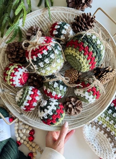 A hand holds a tray of crocheted Christmas ornaments, including a charming red, green, and white crochet stocking, surrounded by pinecones.