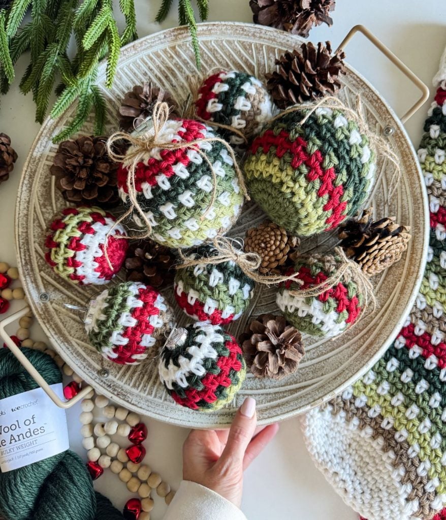 A hand holds a tray of crocheted Christmas ornaments, including a charming red, green, and white crochet stocking, surrounded by pinecones.