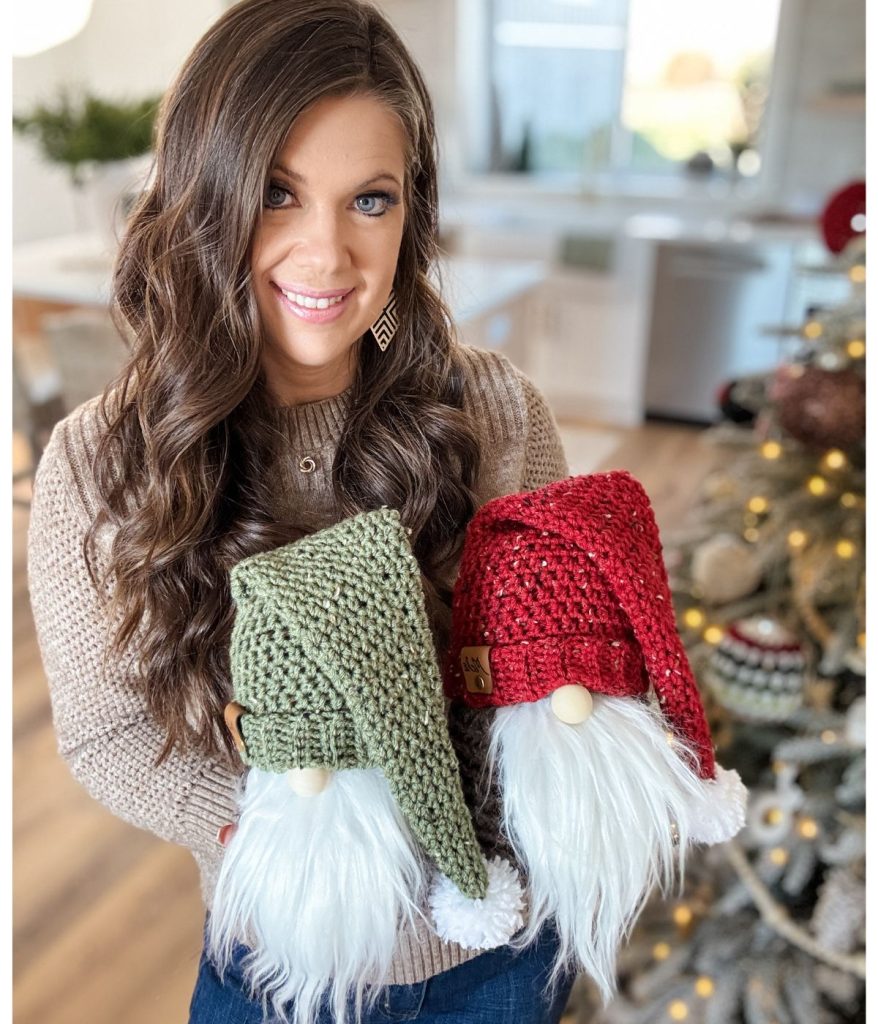 Standing near a beautifully decorated Christmas tree in a bright room, a woman cradles green and red crochet gnome toys with fluffy white beards.