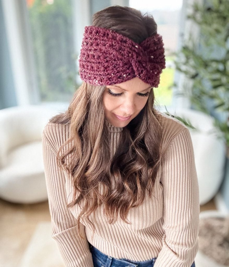 A woman with long hair wearing a burgundy crochet headband and a beige sweater stands in a room with a white chair and plants in the background.