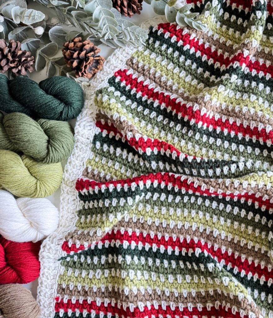 A moss stitch blanket, striped in red, green, white, and beige, is displayed with matching yarn skeins and pinecones nestled among festive greenery.
