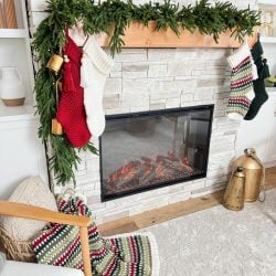 A cozy fireplace with a white stone surround showcases four knitted stockings and a beautifully intricate crochet stocking. Decorated with greenery, it captures the essence of holiday weave. Nearby, a crocheted blanket drapes gracefully over a chair.