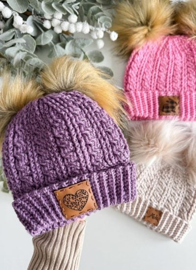 Three cozy crochet beanies with faux fur pom-poms in purple, pink, and white are artfully displayed on a white surface, accented by delicate eucalyptus leaves.